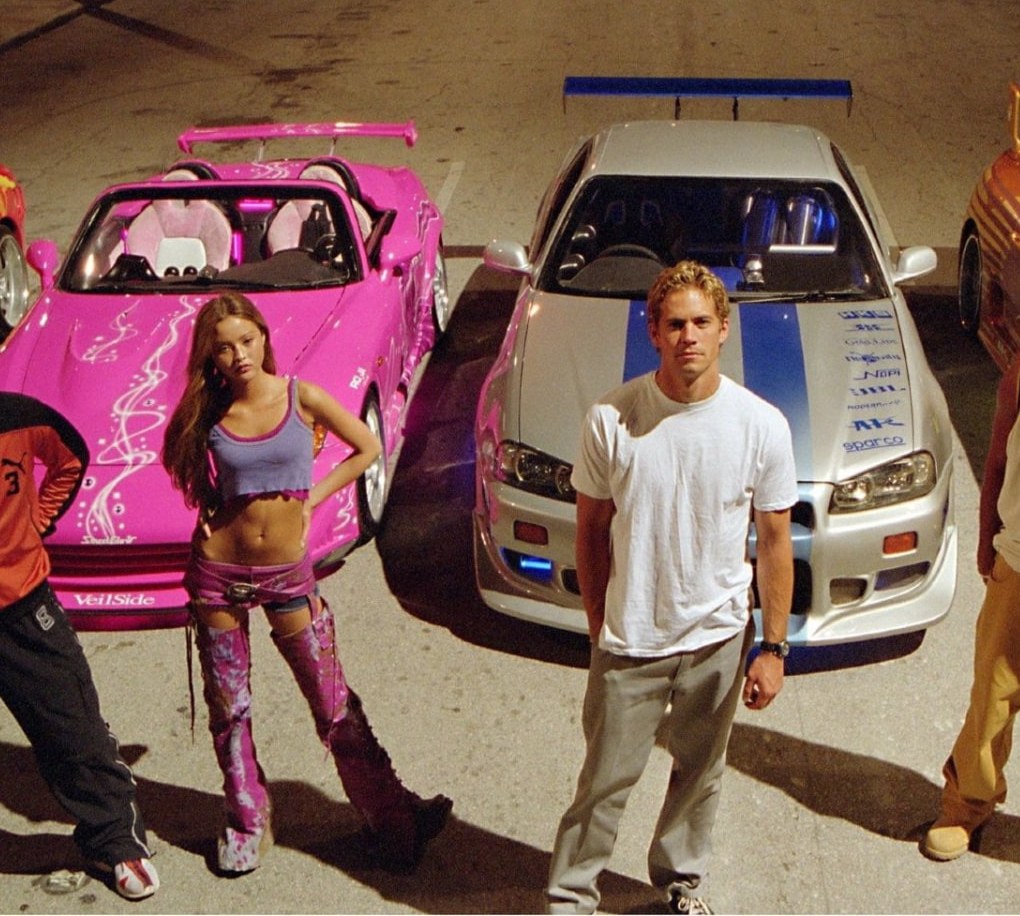 2 Fast 2 Furious cast with the orange Toyota Supra, silver Nissan Skyline GT-R R34, pink Honda Eclipse and red Mitsubishi Lancer Evolution on a neon-lit street at night